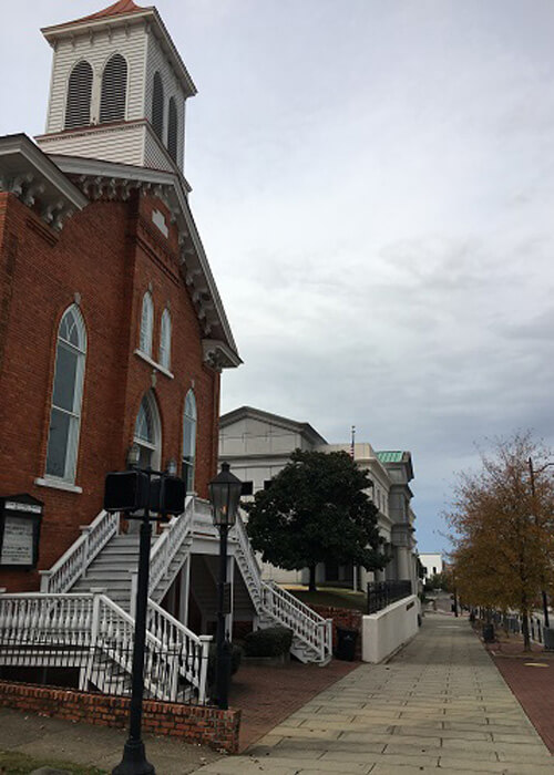 Brick church at left with white wooden tower perched on top, sidewalk leading straight ahead past some other buildings. Grey overcast sky.