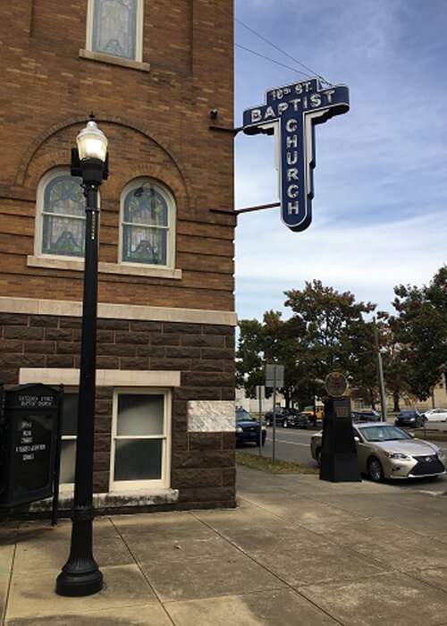 Brick building at left with neon sign hanging from corner reading "16th Street Baptist Church."  Hazy blue sky in background.