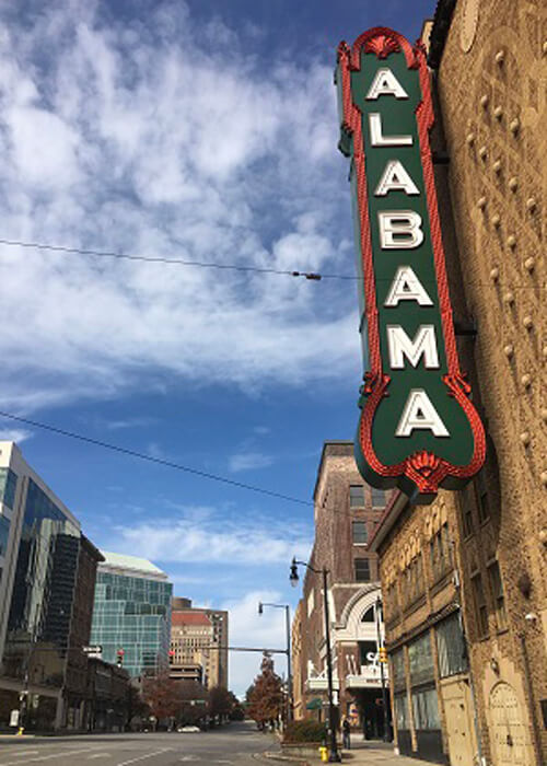 Two-story high 1920's green theatre sign with white lettering reading "Alabama" with downtown Birmingham buildings in the background.  Blue sky with scattered clouds.