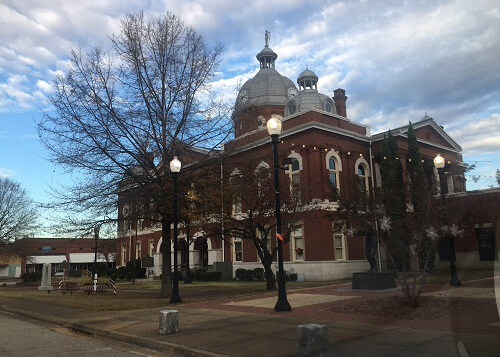 Two-story brick courthouse building with round tower on top and 1950's style streetlamps lit in front.  Blue sky with some clouds.