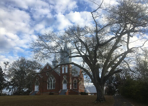 Ornate single-story colonial style brick First Universal Church with white trim in the distance among trees.