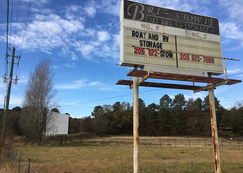 Abandoned drive-in theatre, screen in the distance at left and old drive-in sign at upper right with lettering that reads "boat and RV storage," blue sky with scattered clouds in background.