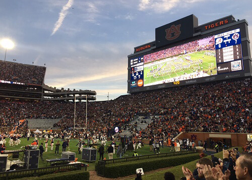 Inside of packed football stadium, video screen of game at upper right, sundown sky with some clouds above.