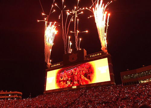 Inside of football stadium at night, fireworks launching above the scoreboard and packed crowd in stands below.