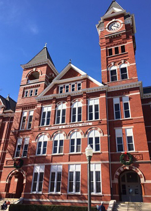 Brick colonial-style three-story campus building at Auburn University, clock tower at right, blue sky in background.