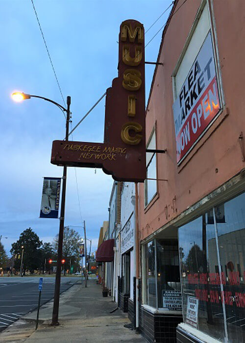 View of downtown Tuskagee with vertical sign reading "music" hanging above orange painted building with other buildings next door.  Light-blue sunrise sky.