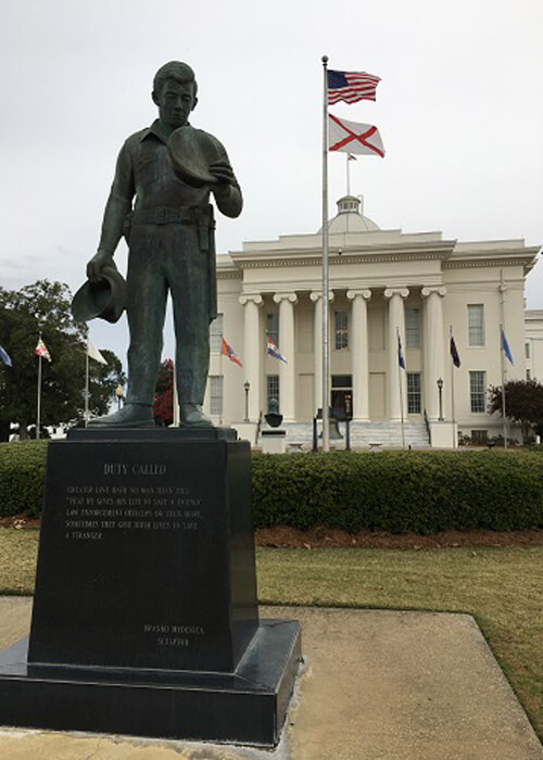 Alabama State Capitol Building, a two-story white classical-style building with greek columns in front.  Statue at left.