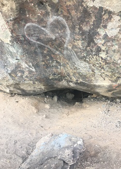 Bottom of giant rock in desert sand, with a small entry opening.  A white heart spray painted above opening.