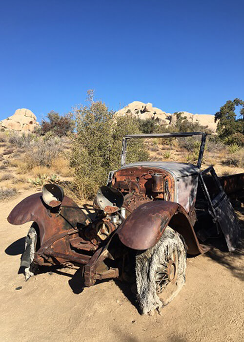 Remnants of old rusty model T ford in the desert dirt, some bushes and rock behind. Blue sky with no clouds.