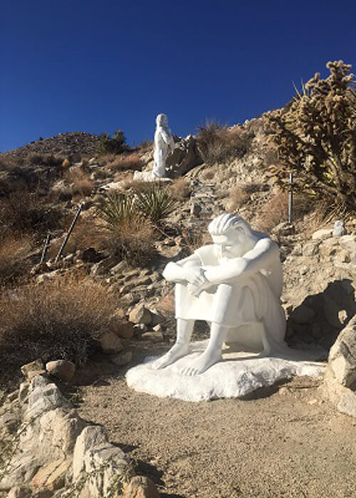 Two white Christian statues at Desert Christ Park sitting amidst the desert terrain of rocks and desert brush.