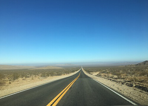 Desert road disappearing into the horizon, desert sand and brush on both sides, blue sky above.