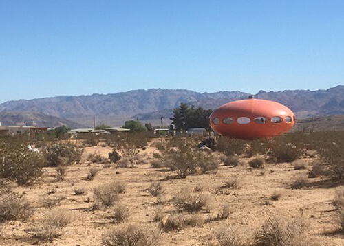 Orange UFO looking structure at right in the desert sand and brush, ridge of mountains in the background and blue sky above.