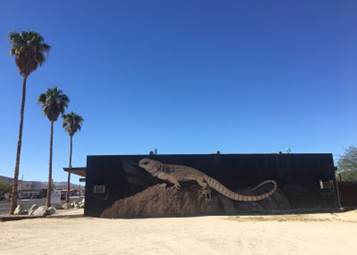 Painting of a giant lizard on side of building, palm trees at left, blue sky with no clouds.