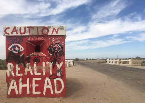 Painted cement barrack that says "caution: reality ahead" in red and white paint.  Road at right,  Blue sky with scattered clouds above