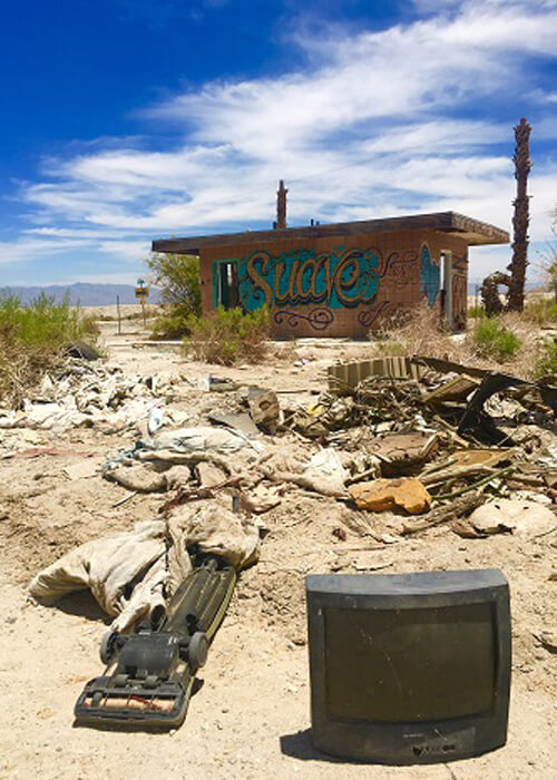 Random junk and old TV on desert ground with an orange cement barrack in background painted with graffiti, blue sky with scattered clouds.