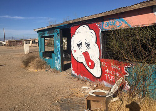 Graffiti painted on side of abandoned cement building, with a white puffy face with red lips and other random art scribbled on building.  Blue sky with no clouds.