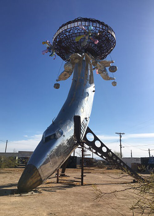 Two-story high aluminum banana shaped art structure, with UFO like elliptical sphere perched on top.  Blue sky with no clouds.