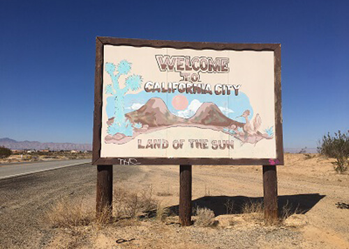 California City welcome sign saying "welcome to California City, land of the sun" at the right of the road leading into town.  Blue sky with no clouds.