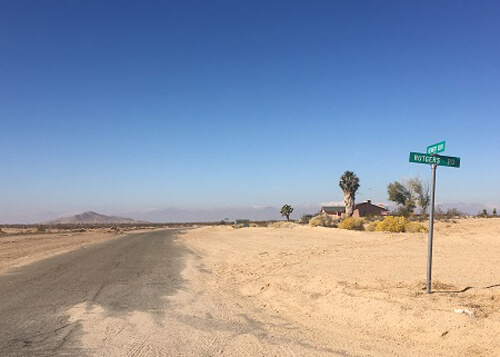 Street sign in middle of desert at right, and street disappearing into nowhere at left.  A couple desert hills in background, blue sky with no clouds.