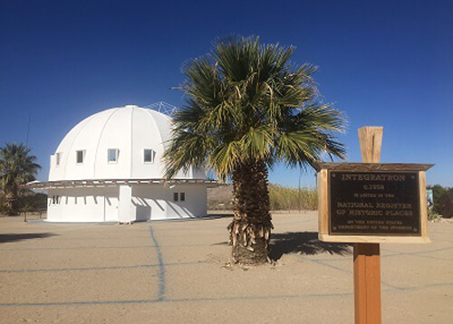 Semi-dome three-story tall white structure at left next to short palm tree in middle and sign at right foreground saying "Integratron".