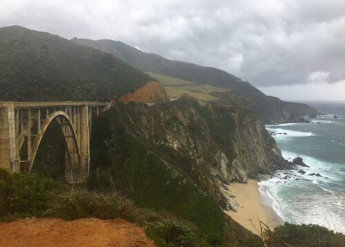 Large cement arched support street bridge at left, with cliffsides at center and ocean to the right. Cloudy skies above.