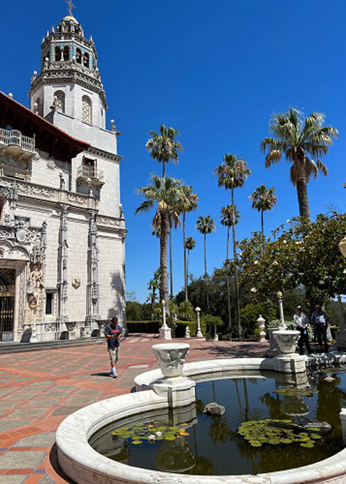 A large Spanish-style tower at left, water fountain and pond at below right, and palm trees at center. Blue sky above. 