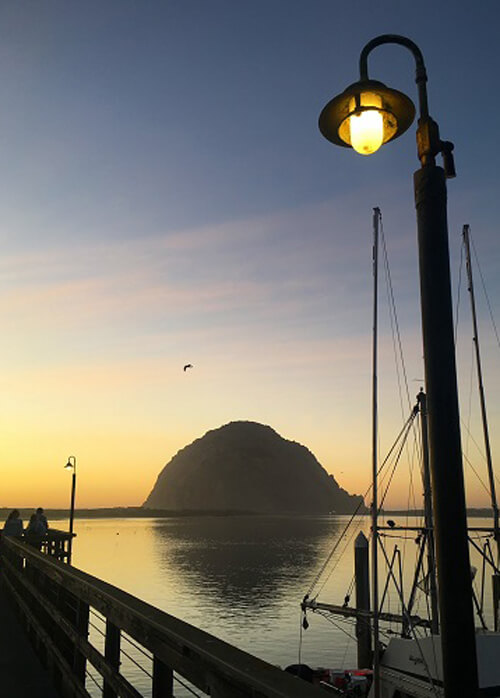 Large rock in the sea at sundown, pier at left and pier light lit at upper right. Rock reflecting in the sea.