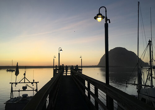 Large rock in the sea at right at sundown, pier at center and pier light lat upper right. Rock reflecting in the sea.