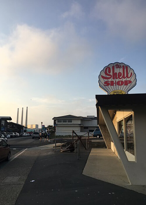 Three power station smokestacks at left in the distance. Shop at right with sign that says "Shell Shop" on the roof. Blue sky with some fog and clouds.