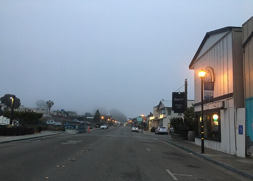 Looking down Main Street of Morro Bay in the morning fog, with some street lights lit and some lights in shop windows. 