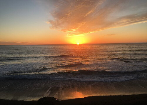 Sunset over the ocean and beach in Cambria, sun almost down and reflecting shades of orange on the ocean and water covered sand.