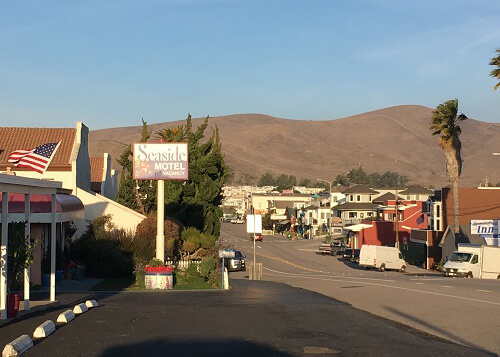 Looking down the main street of small beach town called Cayucos, with single story buildings at left and some homes in distance at the right. Mountains behind the homes, blue sky above.