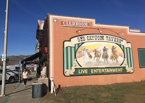 Mural on side of orange building that says "Old Cayucos Tavern," lady walking on sidewalk at left. Blue sky above. 