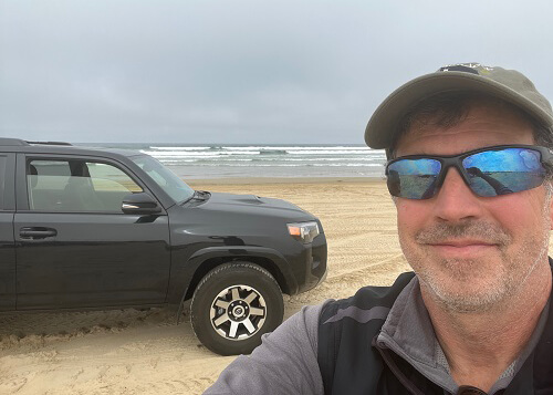 Close up of Mark Loftin at right wearing cap and sunglasses, black Toyota 4Runner car at left on the beach with ocean in background.