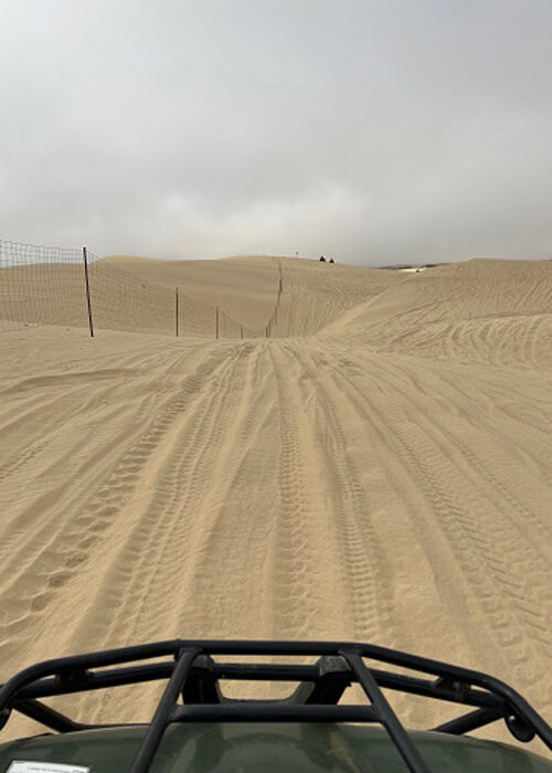 The top portion of hood of sand dune buggy at bottom, rolling sand dunes ahead. Grey sky.