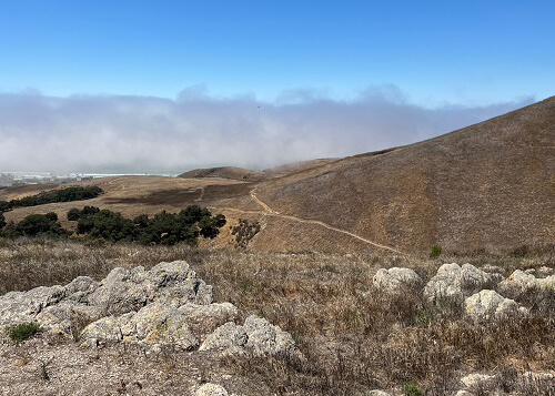 Series of rocks in foreground, with some trails on side of hill in background. Thick layer of fog with blue sky above the fog.