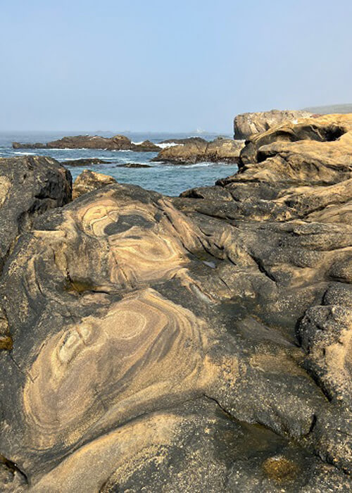 View of ocean with tan and black igneous rock formations in foreground. Blue sky above.