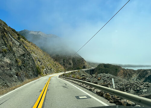 Single-lane road with mountains at left and ocean at right. Fog mist coming in over the road from ocean.