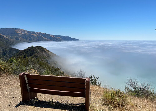 Bench at lower left, overlooking a bed of clouds covering the ocean below. Range of mountains at left and blue sky above.