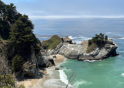 Looking down green water beach cove, steep cliffs at left and waterfall flowing onto the sand below. Blue sky in background. 