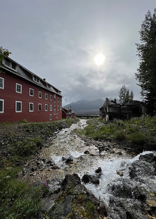 Looking down stream with old red mine processing building on left and old house on right, sun coming through thick grey clouds