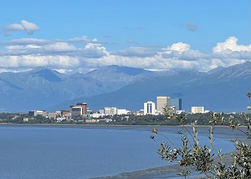 View of downtown Anchorage buildings from across the water, mountain range behind the city, blue sky with clouds above.