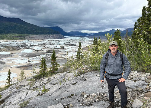 Mark Loftin at right on a trail with lake at left filled with ice chunks, mountains in the distance and cloudy sky above