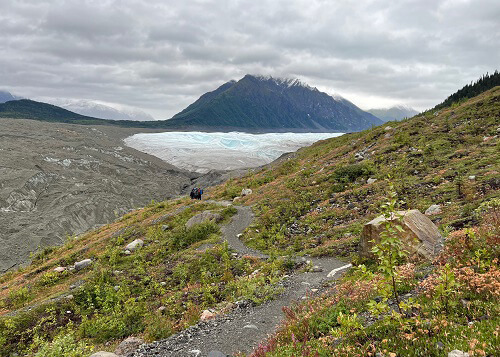 Hiking path winding around hill with glacier ice in the distance, cloudy sky above