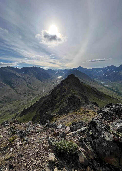 View from mountain top looking down on a series of rocky mountain ridges and river in the far distance, blue hazy sky above.