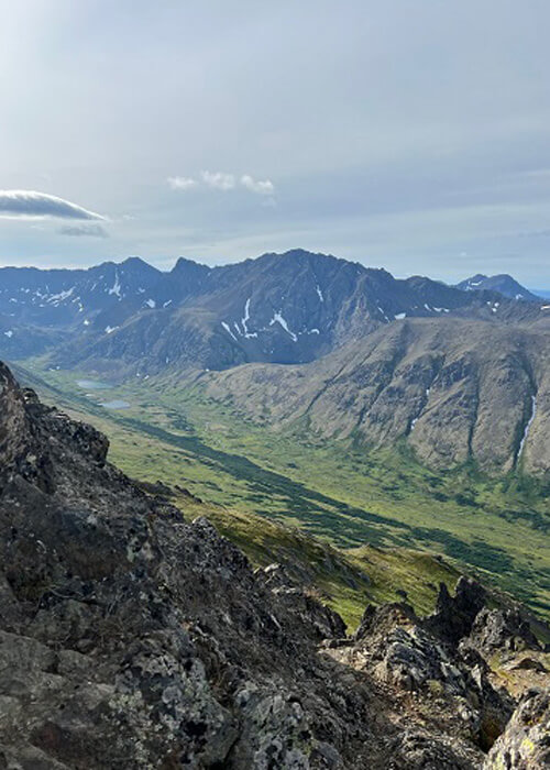 View of ridge of mountains with patch of green in valley below, blue sky with some clouds above.