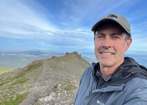 Close up of Mark Loftin with downward sloping hill behind and Anchorage in the far distance, blue sky above.