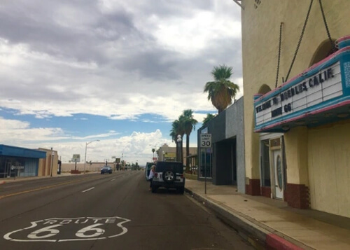 Route 66 sign painted in road at bottom left, with old theatre marquee at right, followed by some palm trees and cloud covered sky in the background.