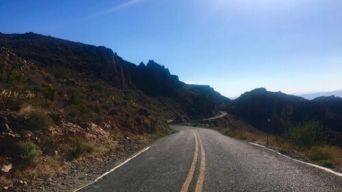 Two lane road twisting into dark brown rocky desert hills, with blue sky in the background.