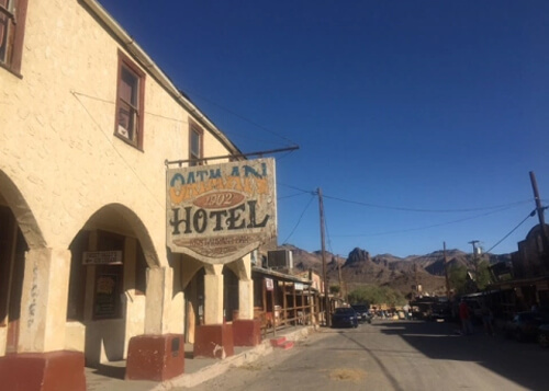 Beige two-story hotel at left, with street disappearing into the mountains. Hotel displays sign saying "Oatman Hotel, 1902." Blue sky in background.